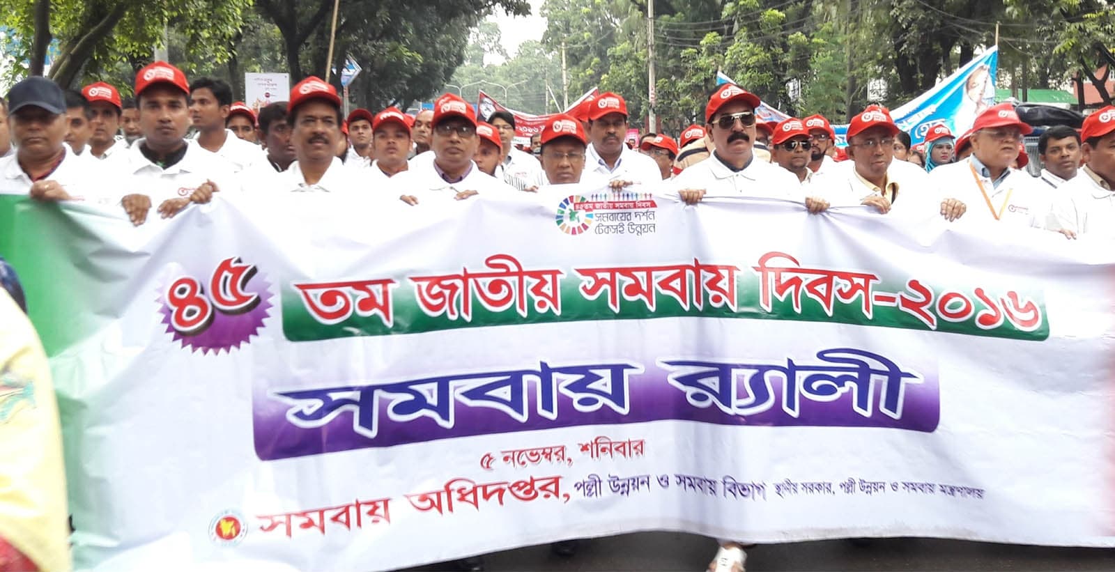 Cooperative members in matching white shirts and red caps carry a wide banner through Dhaka for Bangladesh's 45th National Cooperative Day in November 2016, the Bengali lettering across it reading 'Cooperative Rally'