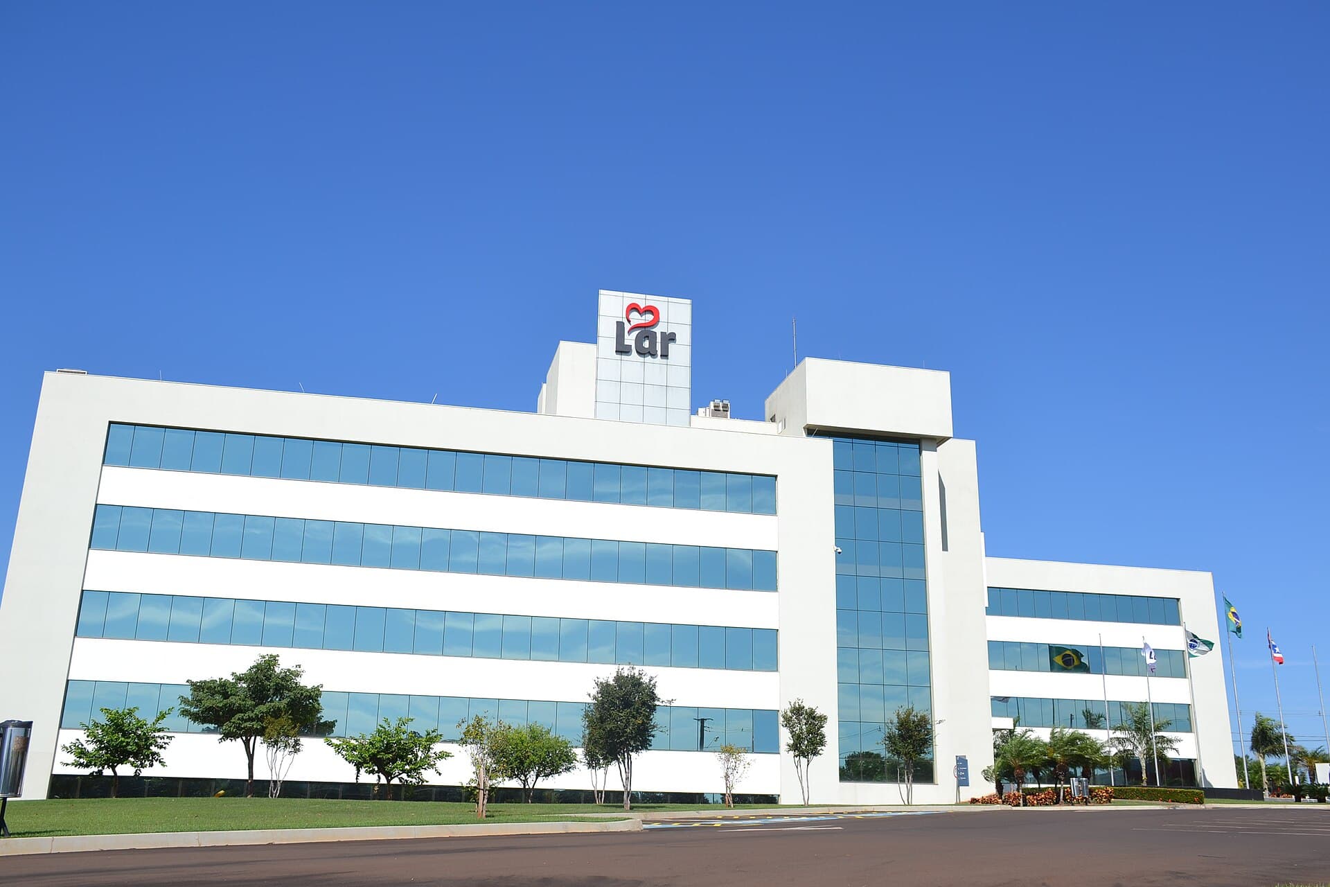 The white glass-fronted headquarters of Lar Cooperativa Agroindustrial, a Brazilian agro-industrial cooperative, its rooftop sign standing against a cloudless blue sky with the Brazilian flag and others flying at the entrance