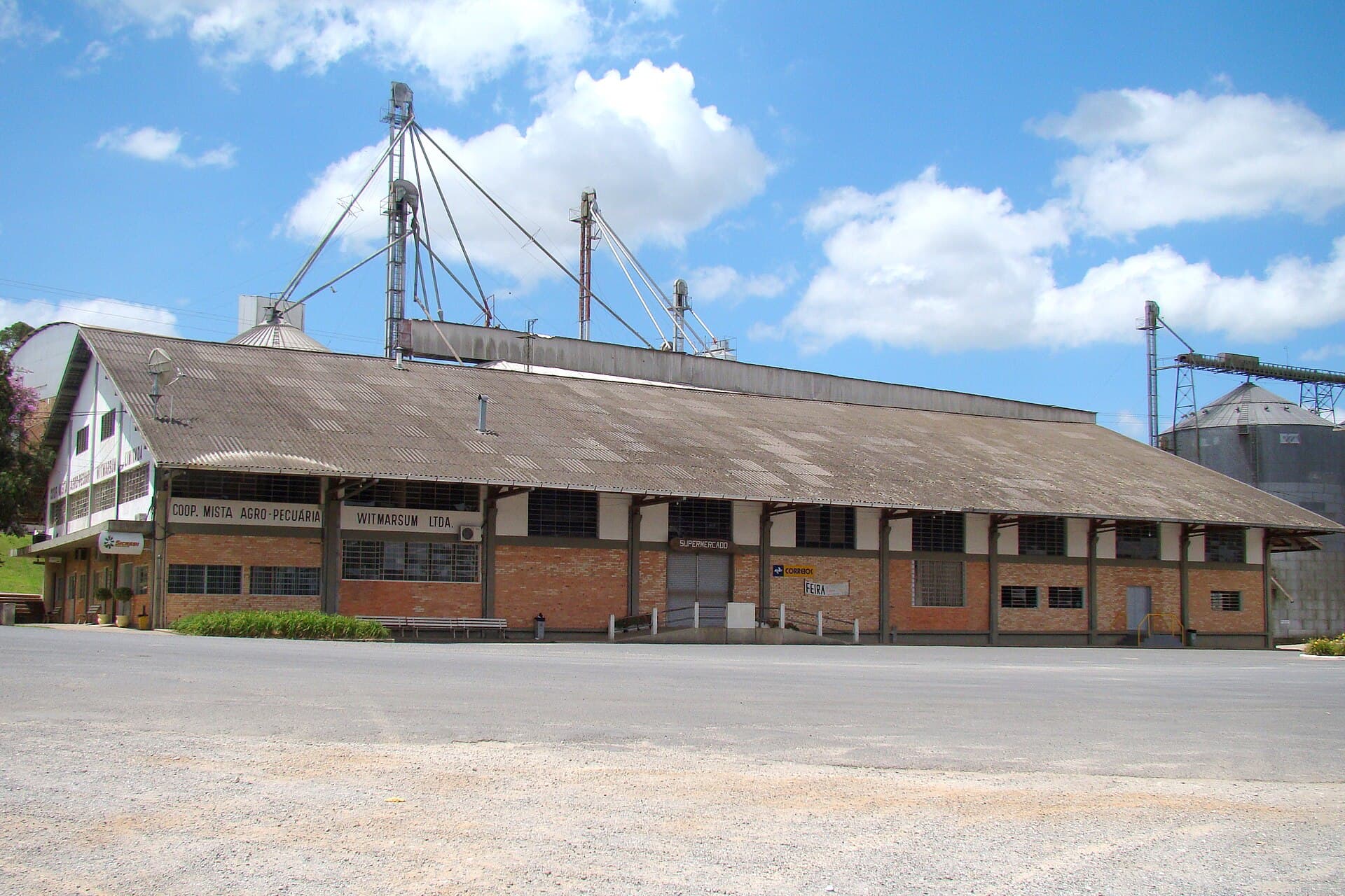 The long tiled-roof premises of Cooperativa Mista Agro-Pecuária Witmarsum at Colônia Witmarsum in Paraná, southern Brazil — the cooperative's name lettered along the brick facade above its member supermarket and produce market, with grain silos and elevator towers rising behind the roofline
