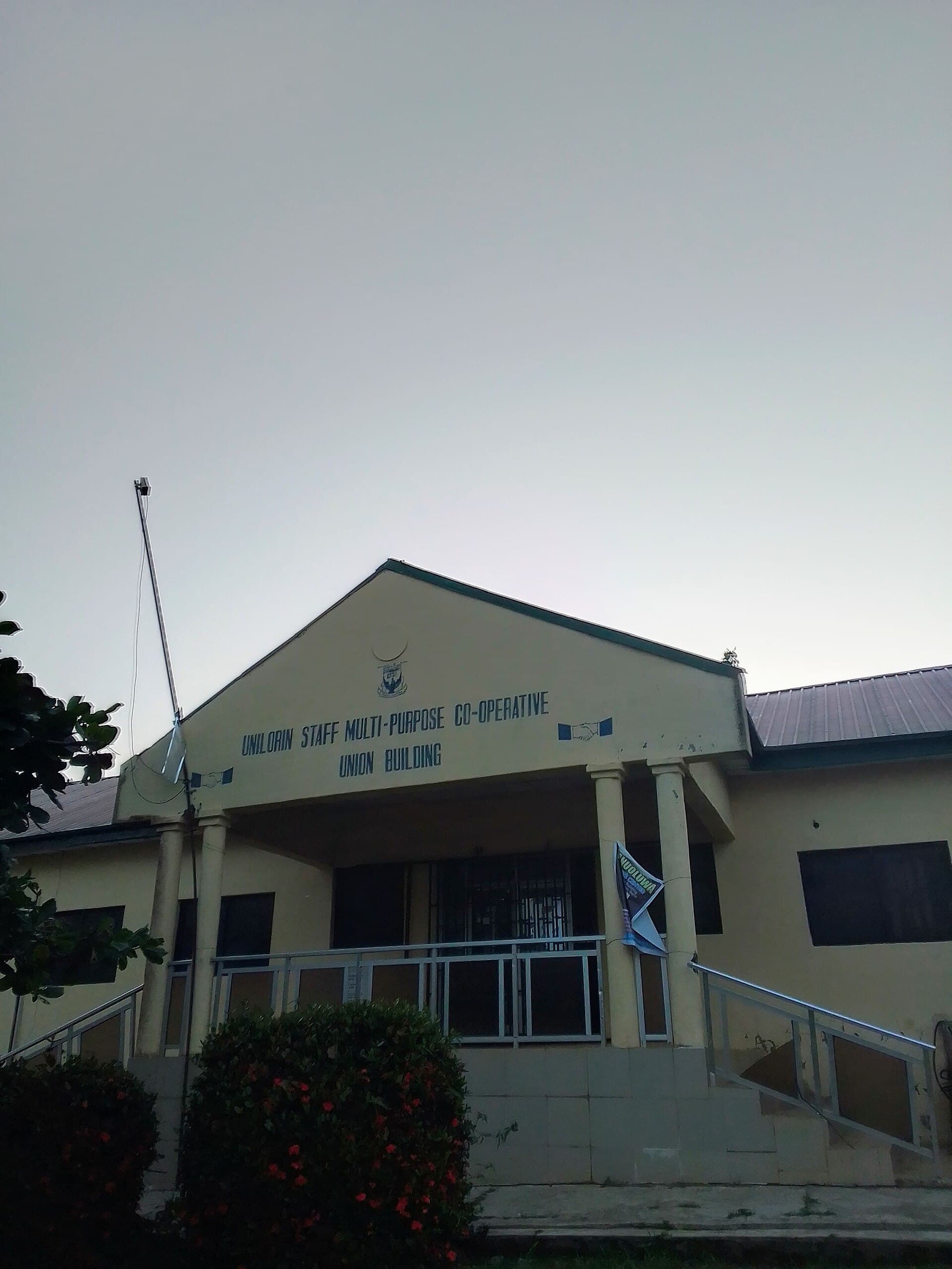 The Unilorin Staff Multi-Purpose Co-operative Union building at the University of Ilorin, Nigeria, at dusk — a pale block with a pillared porch and metal railings, the cooperative's name and two clasped-hands emblems lettered across the gable