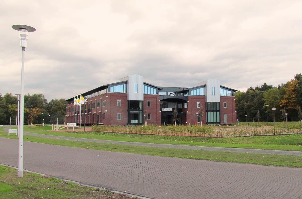 The brick-and-glass headquarters of Agrifirm, a Dutch farmers' agricultural and horticultural cooperative, at Apeldoorn in the Netherlands — a modern two-storey office with twin clad towers, the Agrifirm flag and sign at the entrance across a landscaped forecourt