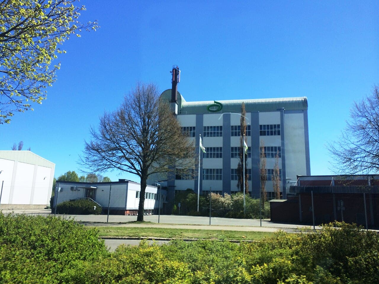 Tall grain silos operated by Lantmännen, the Swedish farmers' agricultural cooperative, at Skara in central Sweden — a pale multi-storey elevator building carrying the cooperative's green roundel logo, seen across a fenced yard on a clear spring day