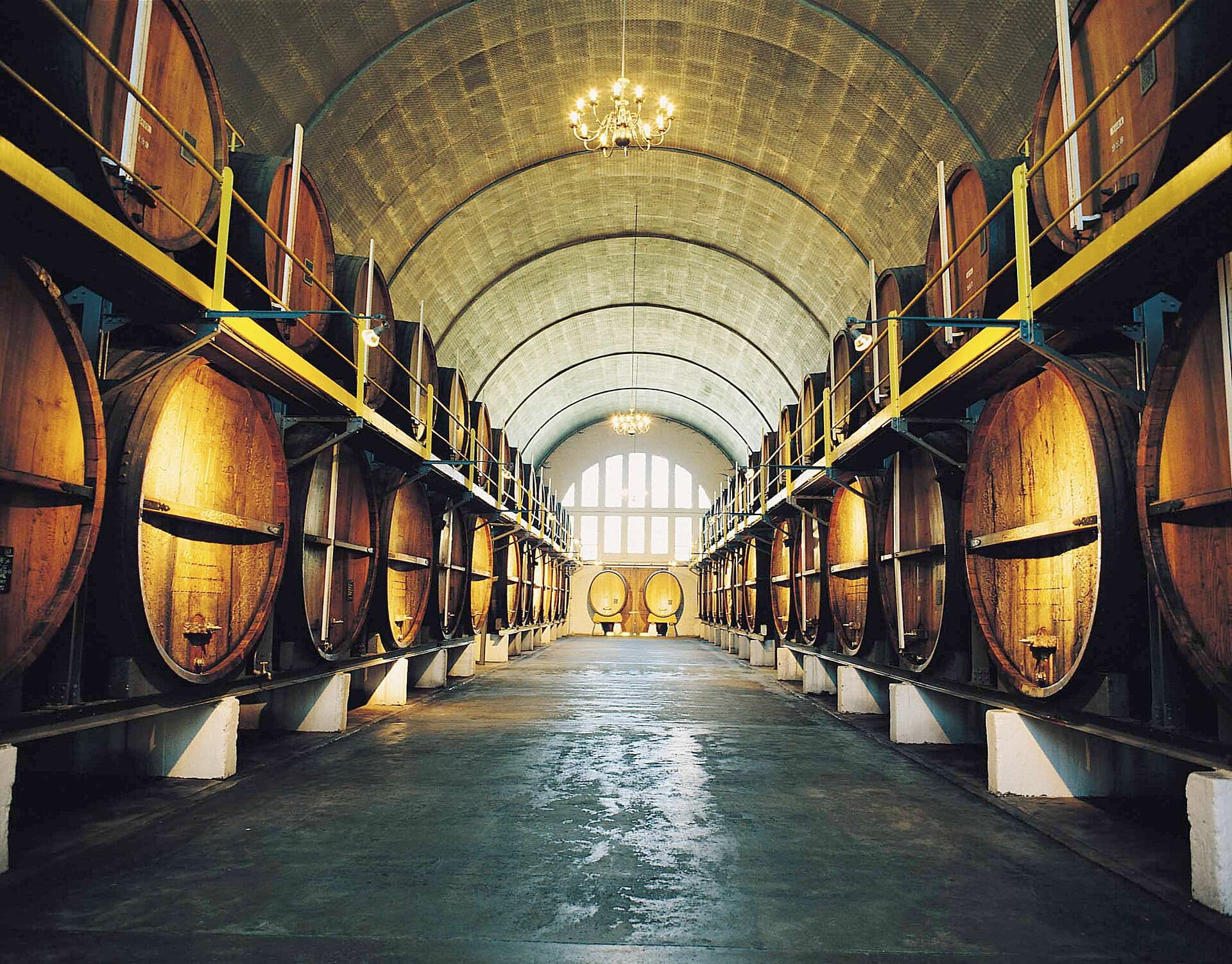 Inside KWV's Cathedral Cellar in South Africa, the maturation hall of a business founded in 1918 as a co-operative of the country's wine growers — a long barrel-vaulted nave lined both sides with towering oak foudres on yellow-railed gantries, chandeliers hanging the length of the aisle towards an arched window