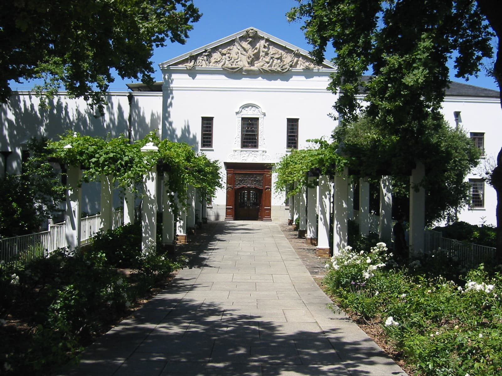 The white gabled headquarters building of KWV at Paarl in the Western Cape — KWV was formed in 1918 as the Koöperatieve Wijnbouwers Vereniging, a co-operative of South African wine growers — seen down a paved path between two pergolas of trained vines and beds of white roses, a sculpted pediment above its carved wooden doors